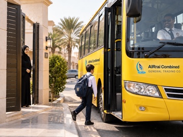 A boy with a backpack walks from his home gate to a waiting yellow AlRrai Combined school bus while his mother watches from the door.